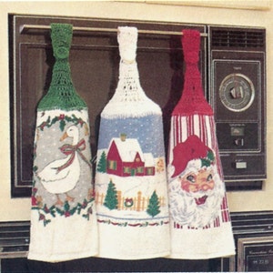 May include: Three crocheted oven mitts hanging on a rack. The mitts are white with different colored crocheted tops. The left mitt has a green top and a goose design. The middle mitt has a white top and a winter scene with a house. The right mitt has a red top and a Santa Claus design.