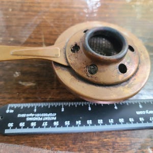 May include: A copper-toned metal object with a handle and a circular top featuring a mesh screen and several holes. A black ruler with white markings is placed next to the object, with the text "PATENT PENDING" visible.