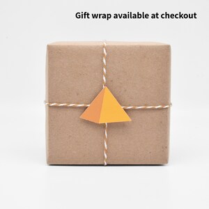 May include: A brown paper-wrapped gift box tied with twine and a small orange and yellow paper pyramid tag. The text "Gift wrap available at checkout" is visible above the box.