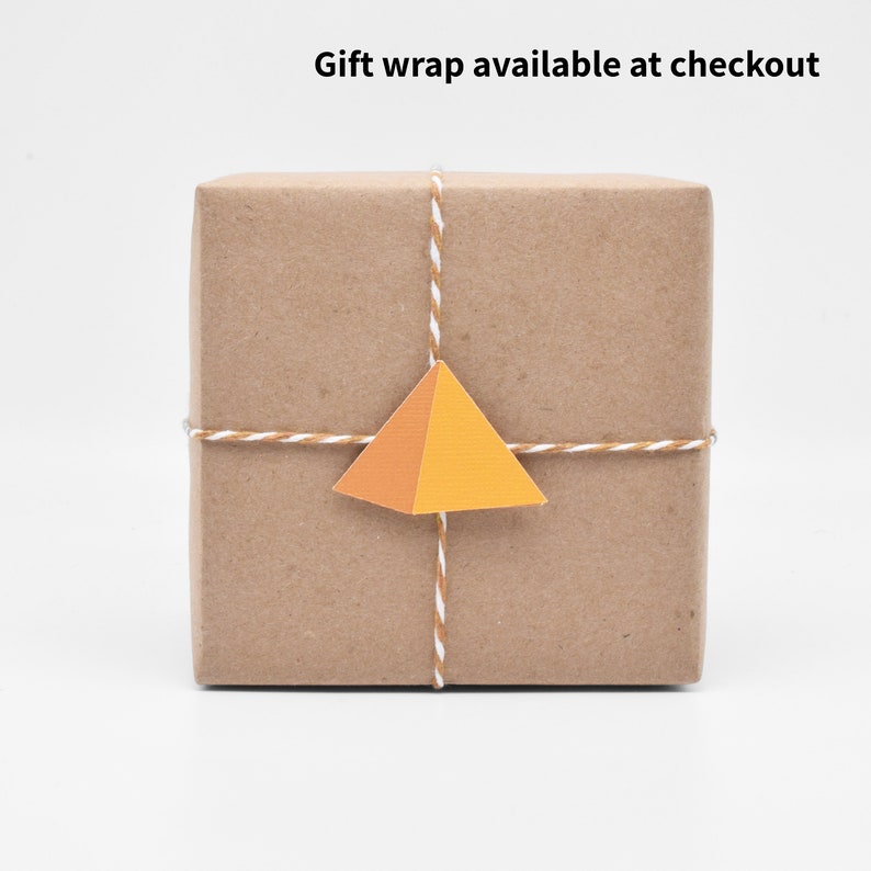 May include: A brown paper-wrapped gift box tied with twine and a small orange and yellow paper pyramid tag. The text "Gift wrap available at checkout" is visible above the box.