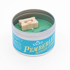 May include: A teal blue candle tin with a white label that reads "Pemberley Scented Candle". The candle features a miniature white building with a green wax landscape.