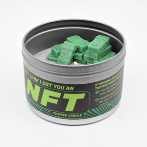 May include: A silver tin candle with a green and black label that reads "HEY LOOK I GOT YOU AN NFT -THEMED CANDLE". The candle is filled with green wax shaped like stacks of money.