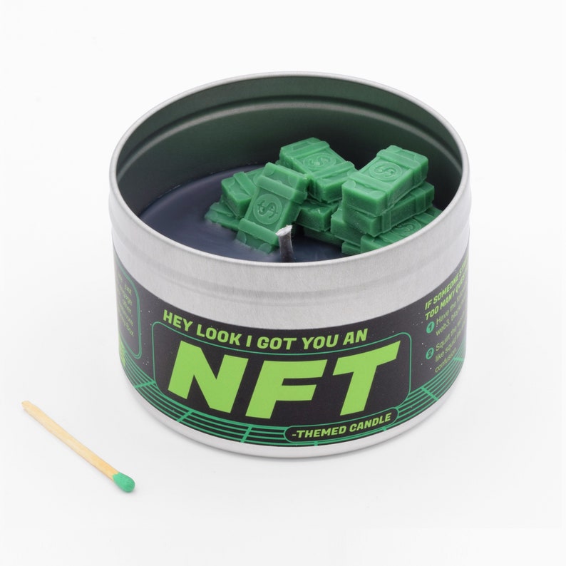 May include: A silver tin candle with a black wax base and green wax stacks shaped like money. The label reads "Hey look I got you an NFT - Themed Candle".