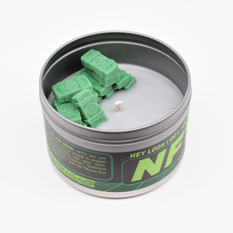 May include: A green and black candle with the text "HEY LOOK I GOT YOU NFT" on the side. The candle is in a metal tin and has a wick sticking out of the top. The candle is surrounded by green wax money stacks.