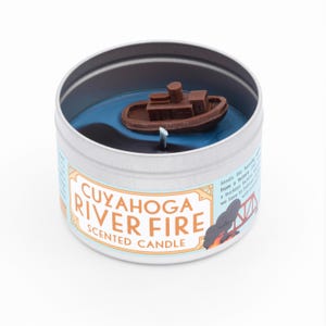 May include: A round, silver tin candle with a blue and black wax design. The candle features a small, brown boat floating on the blue wax. The label reads "CUYAHOGA RIVER FIRE SCENTED CANDLE" and includes a small illustration of a bridge and smoke.