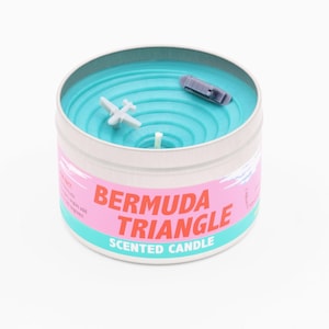 Pode incluir: Uma vela perfumada turquesa em uma lata de prata com um avião e um barco em miniatura no topo. O rótulo diz "Vela perfumada Triângulo das Bermudas".
