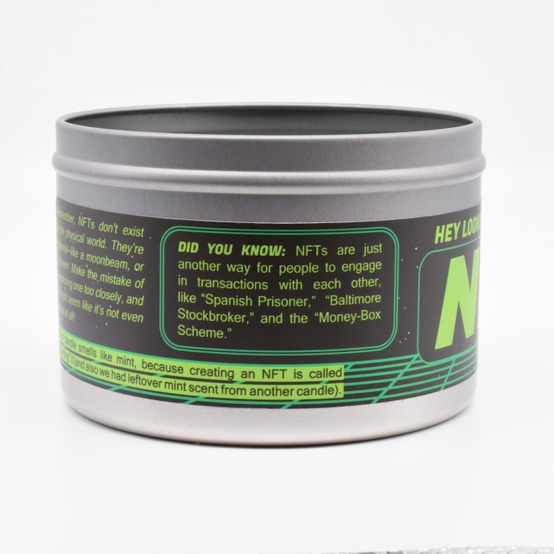 May include: A silver tin candle with a black and green label. The label has the text "HEY LOOK" and "N" in a large font. The text "DID YOU KNOW: NFTs are just another way for people to engage in transactions with each other, like "Spanish Prisoner," "Baltimore Stockbroker," and the "Money-Box Scheme." is also on the label.