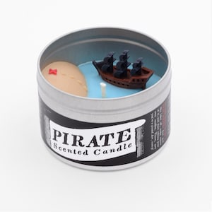 May include: A silver tin candle with a black pirate ship and a small island with a red X on it. The candle is labeled "PIRATE Scented Candle".