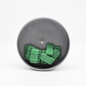 May include: A grey candle jar with a green wax design of stacks of money. The candle is unlit and the wax is still in the jar.