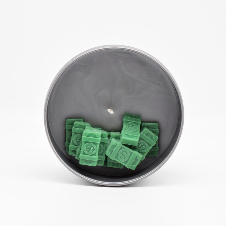 May include: A grey candle jar with a green wax design of stacks of money. The candle is unlit and the wax is still in the jar.
