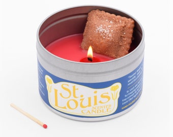Puede incluir: Una vela roja en una lata de plata con un ravioli tostado en la parte superior. La lata tiene una etiqueta azul con el texto "St. Louis Scented Candle" en amarillo.
