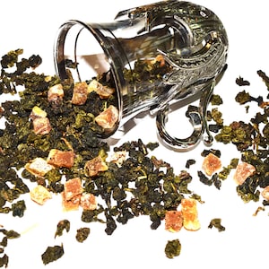 May include: A silver teacup with a decorative design is spilling green tea leaves and dried fruit pieces onto a white surface.