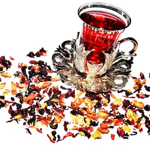 May include: A glass teacup filled with red liquid, resting in an ornate silver-colored holder. Dried tea leaves and fruit are scattered around the cup on a white surface. The cup has a decorative rim and handle.