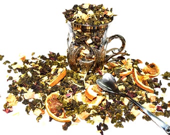 May include: A clear glass cup filled with loose-leaf tea, surrounded by scattered tea leaves, dried orange slices, and a silver spoon. The tea appears to be a blend of green tea with dried fruits and flowers, perfect for a relaxing tea time.