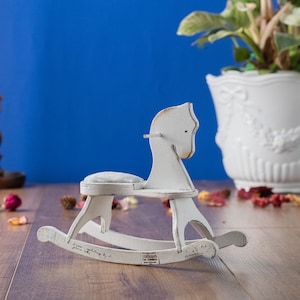 Könnte beinhalten: Ein kleines, weißes Schaukelpferd aus Holz mit einem Used-Look. Das Pferd hat einen grauen Sitz und steht vor einem blauen Hintergrund. Das Schaukelpferd ist ein dekorativer Artikel, möglicherweise für ein Kinderzimmer.