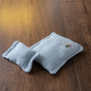 Könnte beinhalten: Zwei hellblaue Leinenkissen auf einer Holzoberfläche. Das größere Kissen hat eine kleine Blumenstickerei in Rosa und Weiß. Das kleinere Kissen ist rechteckig. Beide Kissen haben einen genähten Rand.