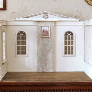 Könnte beinhalten: Ein weißes Miniatur-Puppenhaus mit gewölbten Fenstern und einem kleinen gerahmten Bild. Das Puppenhaus hat einen Holzboden und steht auf einer dunklen Holzoberfläche. Die Wände sind weiß gestrichen und haben eine strukturierte Oberfläche.