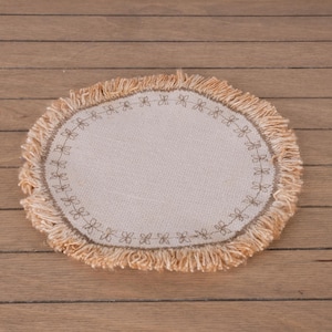 Könnte beinhalten: Ein beigefarbener, ovaler Untersetzer mit Fransen. Die Mitte hat einen gestickten Blumenrand. Der Untersetzer besteht aus einem gewebten Stoff und liegt auf einer Holzoberfläche.