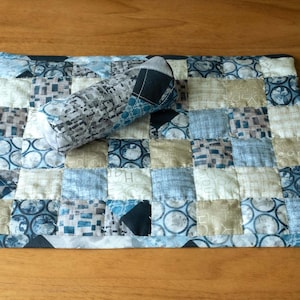 Könnte beinhalten: Ein gesteppter Tischset und ein passender zylindrischer Gegenstand. Der Tischset ist ein Patchwork aus Quadraten in Blau-, Grau- und Beigetönen. Der zylindrische Gegenstand hat ein ähnliches Muster und Farbschema.