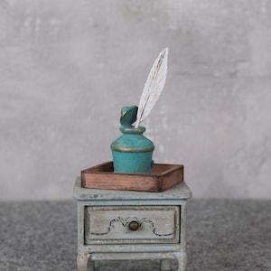 Könnte beinhalten: Ein miniaturer türkisfarbener Tintenfass mit einer weißen Feder, auf einem kleinen Holztablett. Das Tablett steht auf einem antiken, hellblauen Nachttisch mit einer Schublade. Der Tintenfass hat goldene Akzente.