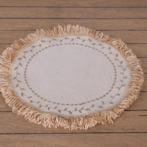 Könnte beinhalten: Ein runder, cremefarbener Platzdeckchen mit Fransenrand. Die Mitte zeigt ein ovales Design mit einem zarten grünen Blättermuster. Das Platzdeckchen besteht aus strukturiertem Stoff und bietet eine rustikale und elegante Ästhetik.