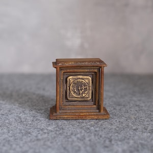 Könnte beinhalten: Eine kleine Uhr im antiken Stil mit quadratischem Zifferblatt und Holzrahmen. Das Zifferblatt ist goldfarben mit schwarzen Zeigern und römischen Ziffern. Die Uhr steht auf einer grauen Oberfläche.