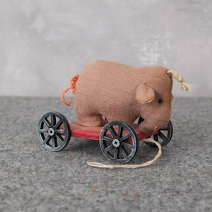 Könnte beinhalten: Ein handgefertigtes Spielzeug-Elefanten aus beigefarbenem Stoff, montiert auf einem roten Holzkarren mit vier schwarzen Speichenrädern. Der Elefant hat kleine Ohren, einen Rüssel und einen Schwanz. Vorne ist eine Schnur befestigt.