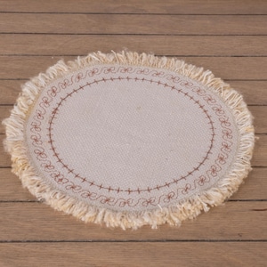 Könnte beinhalten: Ovaler beigefarbener Untersetzer mit Fransenrand und braunem, gesticktem Blumendesign. Der Untersetzer ist aus einem gewebten Stoff gefertigt und liegt auf einer Holzoberfläche. Ein dekorativer Artikel für das Zuhause.