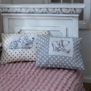 Könnte beinhalten: Zwei Miniaturkissen mit Cartoon-Illustrationen liegen auf einem rosa Bett. Ein Kissen ist beige mit schwarzen Punkten und einer Katzenzeichnung. Das andere ist grau mit weißen Punkten und einer Bärenzeichnung.
