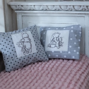 Könnte beinhalten: Zwei Miniatur-Kissen mit grauen Tupfen und Winnie-Puuh- und Ferkel-Stickerei. Die quadratischen Kissen liegen auf einer rosa strukturierten Oberfläche, mit einem weißen Fenster und einem dekorativen Kopfteil im Hintergrund.