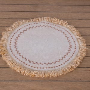 Könnte beinhalten: Ein runder, cremefarbener Platzdeckchen mit einer dekorativen, genähten Bordüre in einem rötlich-braunen Farbton. Der Rand ist mit einem hellbraunen Material gefranst. Auf einer Holzoberfläche platziert.