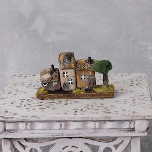 Könnte beinhalten: Eine Miniatur-Dorfszene mit mehreren kleinen, rustikal aussehenden Häusern mit Schornsteinen und Fenstern. Ein kleiner grüner Baum steht neben den Häusern. Die Häuser stehen auf einer rechteckigen Basis mit strukturiertem, braunem Aussehen. Die Szene wird auf einem weißen, verzierten Tisch präsentiert.