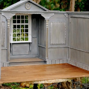 Könnte beinhalten: Ein Miniatur-Holzhaus in Grau mit einem weißen Fenster, dekorativen Säulen und einem Holzboden. Das Haus hat eine kleine Veranda und befindet sich im Freien mit einem natürlichen Hintergrund.