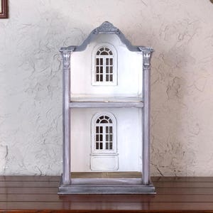 Könnte beinhalten: Ein zweistöckiges Puppenhaus mit weißer Fassade und gewölbten Fenstern. Das Puppenhaus hat eine graue Verkleidung und steht auf einer Holzoberfläche. Die Fenster haben weiße Rahmen und sind in kleine Scheiben unterteilt.