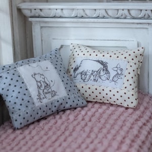 Könnte beinhalten: Zwei dekorative Miniaturkissen mit Polka Dots und gestickten Winnie-the-Pooh-Figuren. Ein Kissen ist hellblau mit dunkelblauen Punkten, das andere cremefarben mit braunen Punkten. Sie liegen auf einer rosa strukturierten Oberfläche.