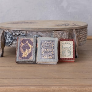 Könnte beinhalten: Drei Miniatur-Antiquitätenbücher mit dekorativen Einbänden, angeordnet auf einer Holzoberfläche. Die Bücher haben verzierte goldene Schriftzüge und Illustrationen. Ein geschnitzter Holzständer befindet sich im Hintergrund.