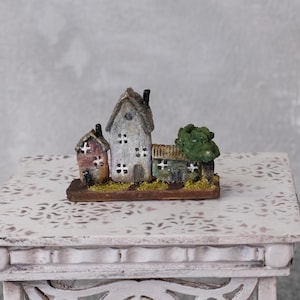 Könnte beinhalten: Eine Miniatur-Dorfszene mit drei rustikalen Häusern in verschiedenen Farben und Texturen. Die Häuser stehen auf einem Holzsockel mit einem kleinen grünen Baum. Die Szene wird auf einem weißen, verzierten Tisch präsentiert.