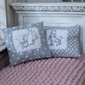 Könnte beinhalten: Zwei dekorative Miniaturkissen mit grauen Polka-Dots und gestickten Zeichentrickfiguren. Ein Kissen zeigt Ferkel, das andere Winnie Puuh. Sie liegen auf einer rosa strukturierten Decke, was auf ein Kinderzimmer oder eine Kindertagesstätte hindeutet.