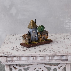 Könnte beinhalten: Eine Miniatur-Dorfszene mit drei kleinen Häusern in Blau, Beige und Grün. Die Häuser stehen auf einer braunen Basis mit einem kleinen grünen Baum. Die Szene wird auf einem weißen, verzierten Tisch präsentiert. Der Tisch ist etwa 60 cm breit.