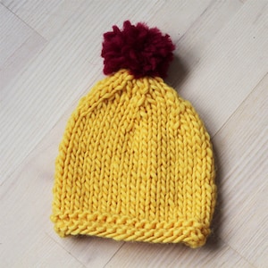 Puede incluir: Un gorro de punto amarillo con un pompón burdeos en la parte superior.