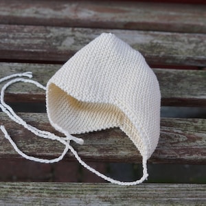 Peut inclure: Un bonnet de bébé en crochet blanc avec une longue attache. Le bonnet est posé sur une surface en bois.