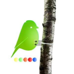 Könnte beinhalten: Eine grüne Vogelsilhouette aus Acryl ist mit einem durchsichtigen Plastikband an einem Baumstamm befestigt. Der Vogel ist nach links gerichtet und hat ein kleines Loch in seinem Körper. Der Baumstamm ist braun und hat eine raue Textur.