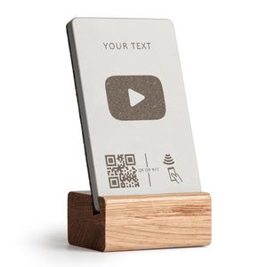Könnte beinhalten: Eine rechteckige, silberfarbene Karte mit dem Text "YOUR TEXT" und einem Wiedergabesymbol, in einem Holzständer. Die Karte enthält auch einen QR-Code und ein NFC-Symbol. Der Ständer ist aus hellem Holz gefertigt.