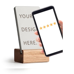 Könnte beinhalten: Ein rechteckiges Schild mit dem Text "YOUR DESIGN HERE" wird auf einem Holzsockel angezeigt. Ein Smartphone mit fünf goldenen Sternen auf dem Bildschirm wird in der Nähe gehalten. Das Schild und das Telefon sind vor weißem Hintergrund.