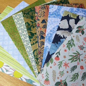 May include: A collection of patterned paper sheets fanned out, showcasing various designs. The papers feature floral, geometric, and abstract patterns in colors like green, blue, gold, and white. The papers are arranged on a light brown wooden surface.