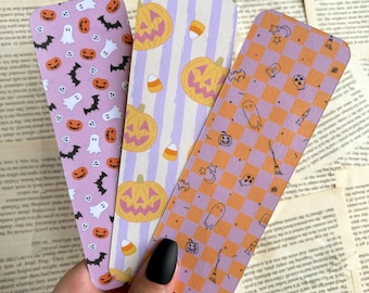 May include: Three Halloween-themed bookmarks with different patterns. One has a lavender background with bats, pumpkins, and ghosts. Another has vertical stripes with pumpkins and candy corn. The third has an orange and lavender checkerboard pattern with Halloween icons.