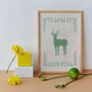 May include: A framed print with a green illustration of a deer with antlers standing in a leafy border on a white background.