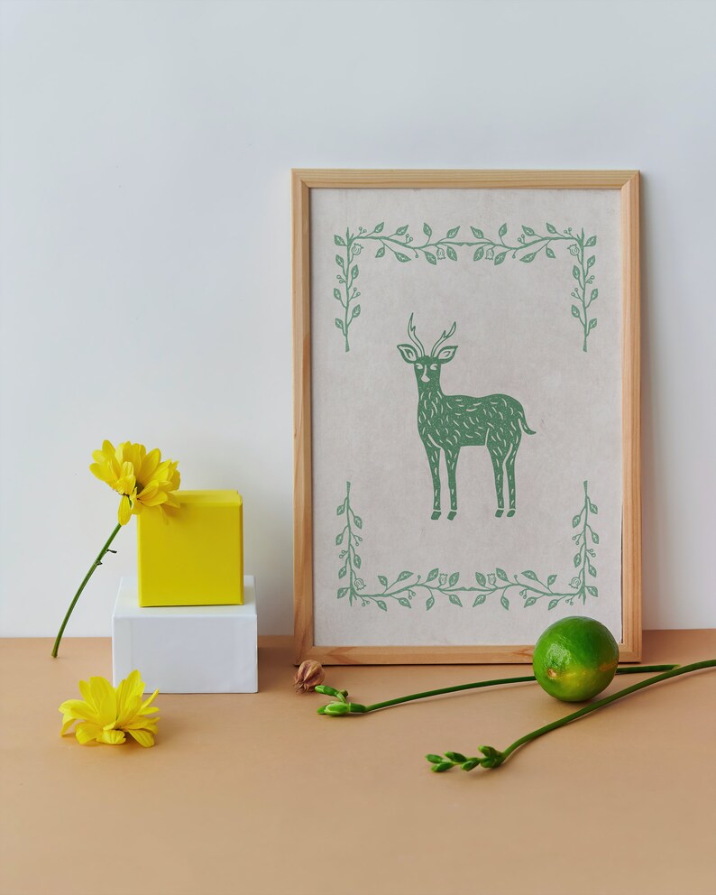 May include: A framed print with a green illustration of a deer with antlers standing in a leafy border on a white background.