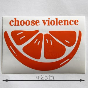 May include: A sticker with the words "choose violence" in red, above a stylized orange slice. The sticker measures 2.9 inches high and 4.25 inches wide.