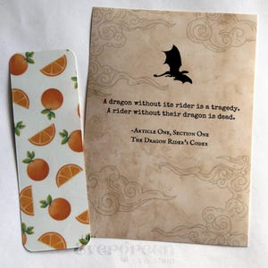 May include: A bookmark with orange slices and a card with a dragon silhouette and text. The bookmark features a white background with orange slices and green leaves. The card has a dragon silhouette and text that reads "A dragon without its rider is a tragedy."